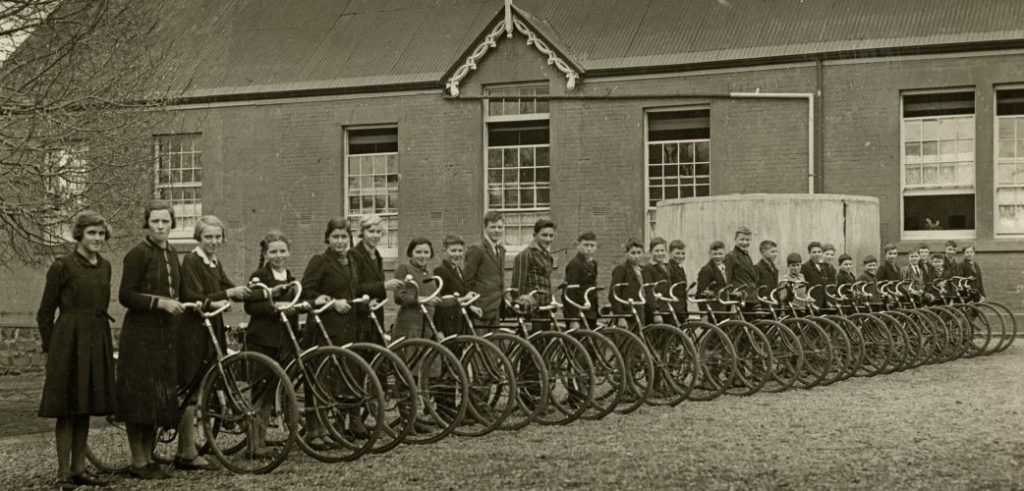 An old black and white photo of a line of school kids and their bikes.