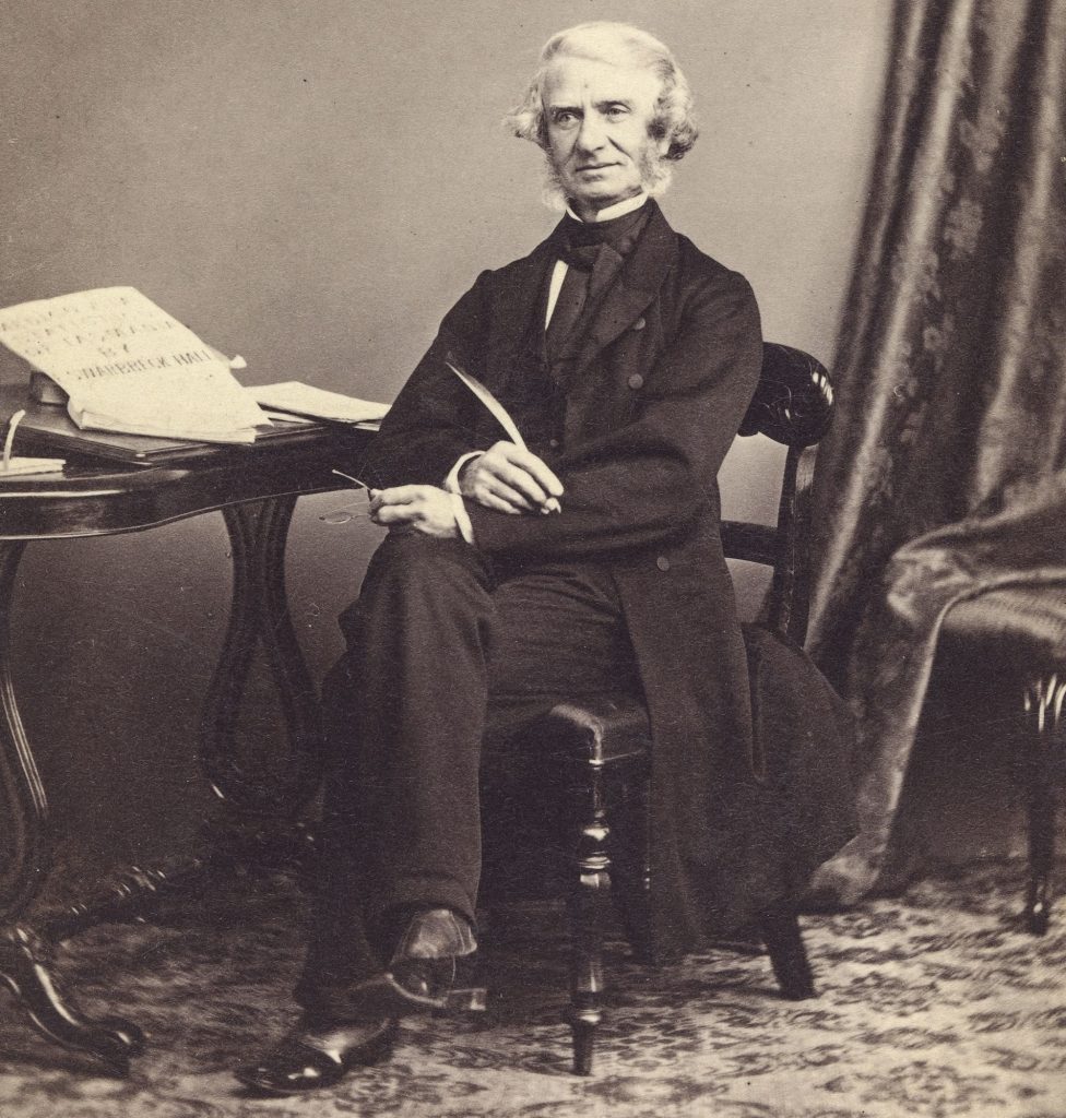 A black and white photo of a man sitting on a char holding a quill and a pair of glasses.