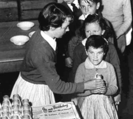 An old black and white photo of children lining up to receive their milk.